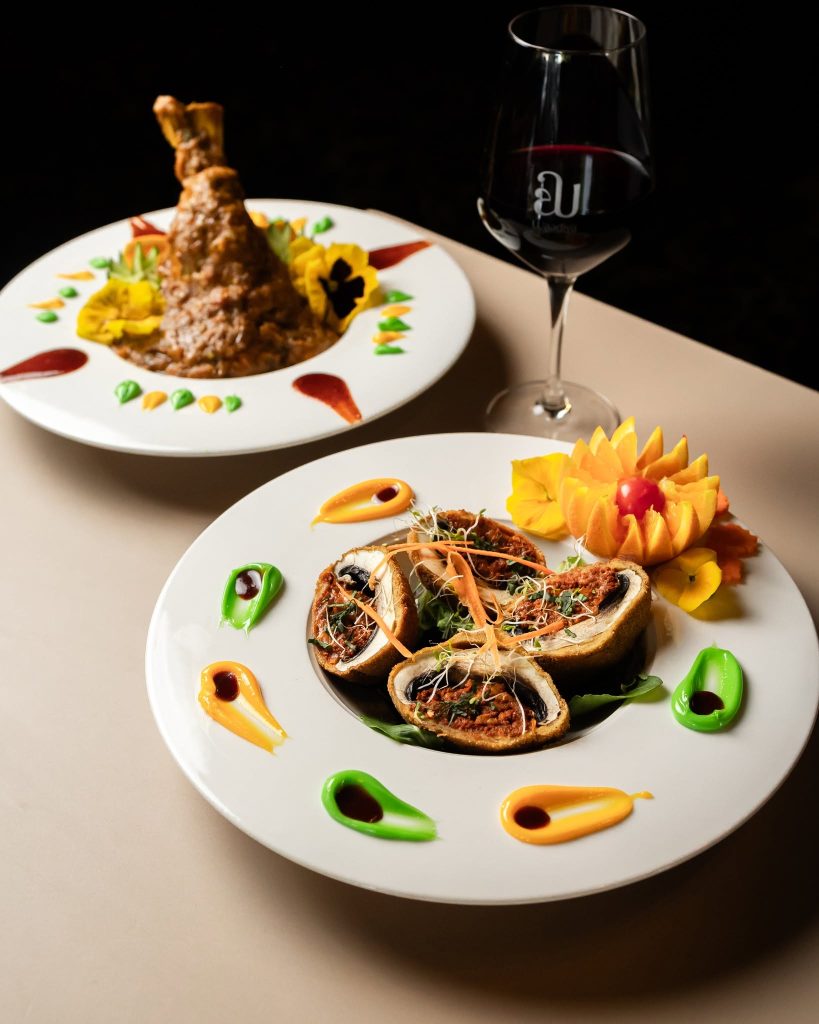 Gourmet plated dishes with roasted meat and stuffed pastries, garnished with colorful sauces and a glass of wine.